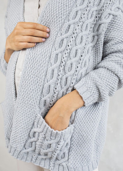 A person wearing a cozy light gray knitted sweater with intricate cable patterns. One hand is in the pocket, conveying warmth and comfort.