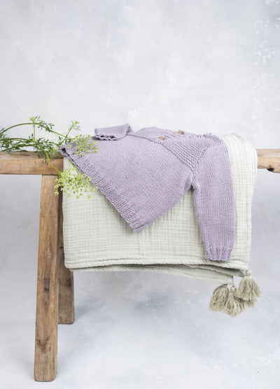A soft, purple knit baby sweater with wooden buttons is neatly draped over a creamy quilt on a rustic wooden bench. Delicate green sprigs add a fresh touch.