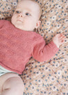 A baby in a pink knitted sweater lies on a floral-patterned blanket, looking to the side with a calm expression. Soft light enhances the serene atmosphere.
