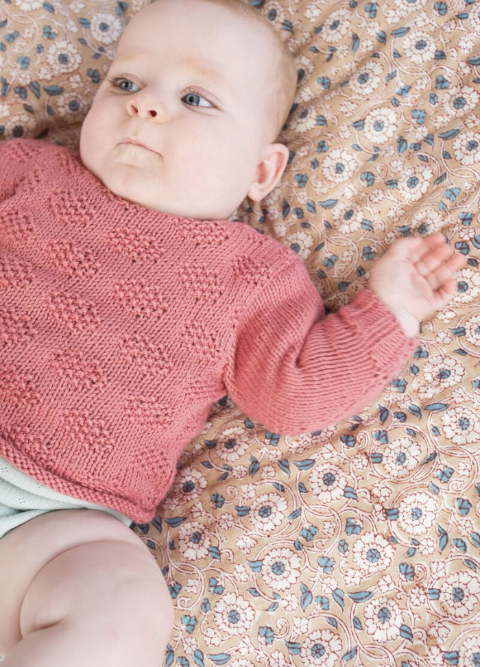 A baby in a pink knitted sweater lies on a floral-patterned blanket, looking to the side with a calm expression. Soft light enhances the serene atmosphere.