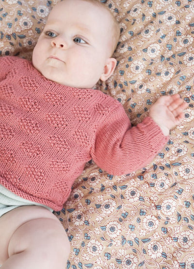 A baby in a pink knitted sweater lies on a floral-patterned blanket, looking to the side with a calm expression. Soft light enhances the serene atmosphere.