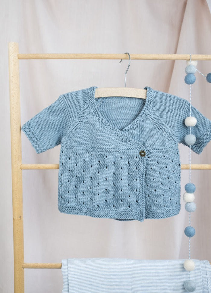 Blue knitted baby cardigan with eyelet pattern hanging on a wooden rack. Soft, cozy ambiance with a backdrop of pastel fabric and felt garland.