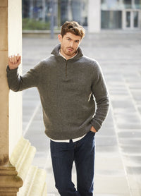 Man with light brown hair leans against a stone wall. He's wearing a gray sweater and jeans, with a casual, thoughtful expression in an urban setting.