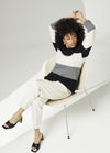 A person sits in a modern chair, their arms raised. They wear a black, gray, and white striped sweater, light pants, and black heels. The scene is minimalist and relaxed.