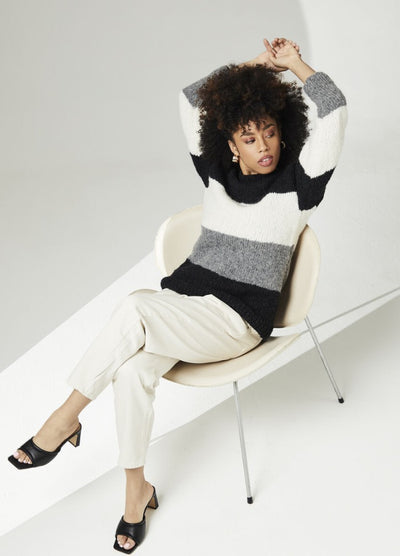 A person sits in a modern chair, their arms raised. They wear a black, gray, and white striped sweater, light pants, and black heels. The scene is minimalist and relaxed.