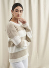 A woman in a beige and white striped sweater and white pants poses against a cream curtain backdrop. She has a thoughtful expression, with hand near chin.