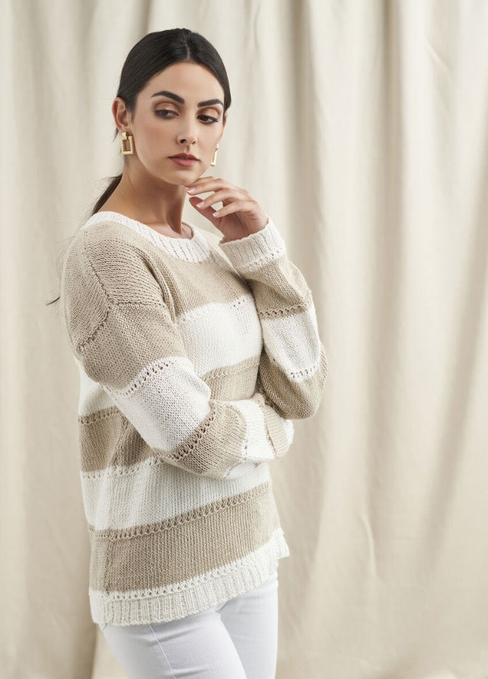 A woman in a beige and white striped sweater and white pants poses against a cream curtain backdrop. She has a thoughtful expression, with hand near chin.