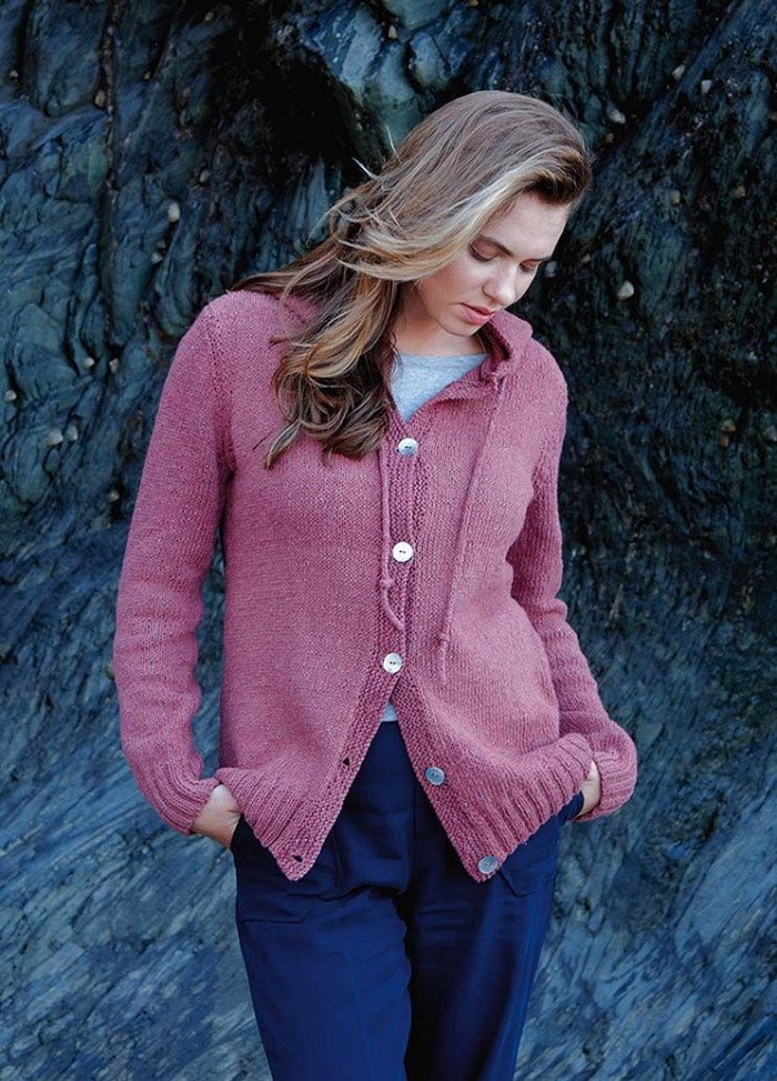 Woman with long hair wearing a pink cardigan and blue pants stands against a textured rock wall, looking down. The scene conveys a calm, introspective mood.