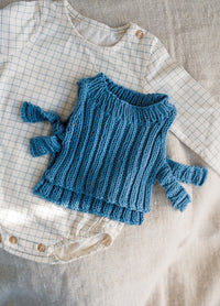 Blue knitted baby sweater on cream checked onesie with wooden buttons, placed on beige fabric. Cozy and soft textures dominate the image.