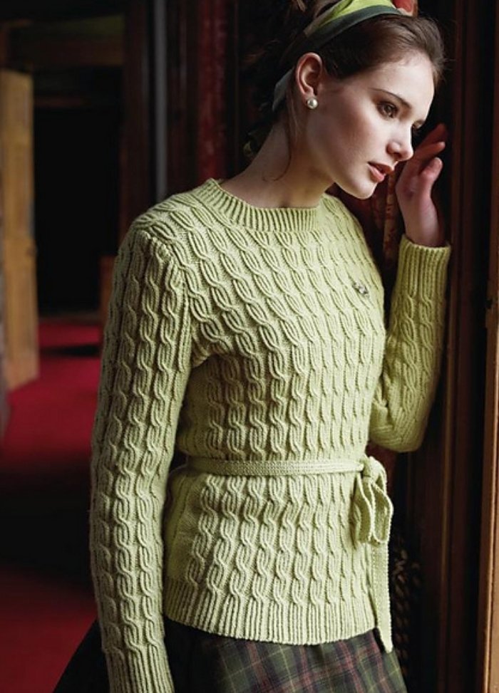 A woman in a green cable-knit sweater and plaid skirt leans against a wooden frame. Her expression is introspective, and the setting is warmly lit.