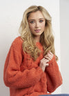 Young woman with long blonde hair wearing a cozy orange sweater, seated with a soft expression against a white background, conveying warmth and comfort.