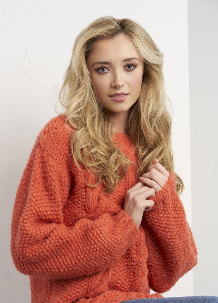 Young woman with long blonde hair wearing a cozy orange sweater, seated with a soft expression against a white background, conveying warmth and comfort.