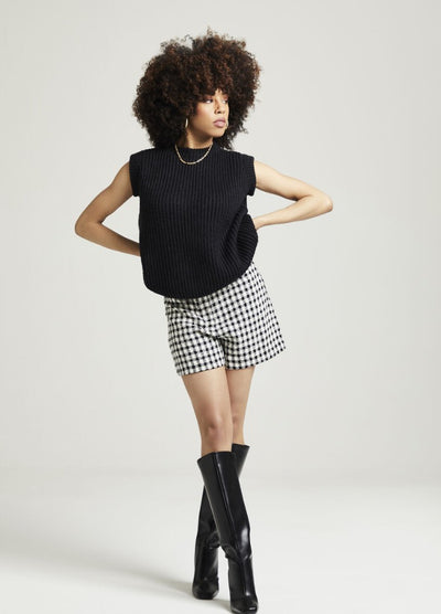 A person with curly hair poses confidently against a plain background. They wear a black sleeveless top, checkered shorts, and knee-high black boots.