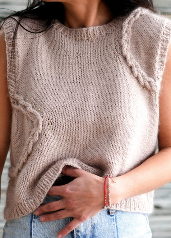 A person wears a handmade beige knitted sweater with cable details, paired with light blue jeans. Their hand rests on their waist, conveying a relaxed, cozy vibe.
