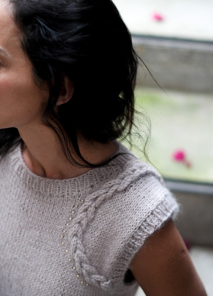 A woman with dark hair faces away, wearing a light gray knit sweater with intricate stitching. The soft lighting and pink flowers create a calm atmosphere.