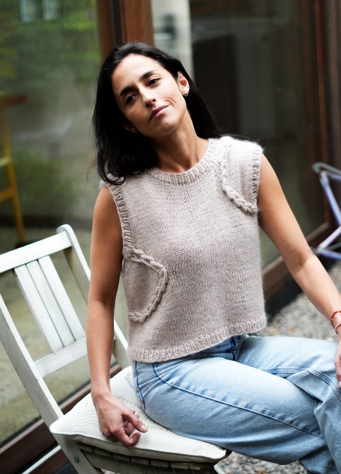 Woman with long dark hair wearing a beige knit sleeveless top and light blue jeans, sitting on a white chair by a window, exuding a relaxed and contemplative vibe.