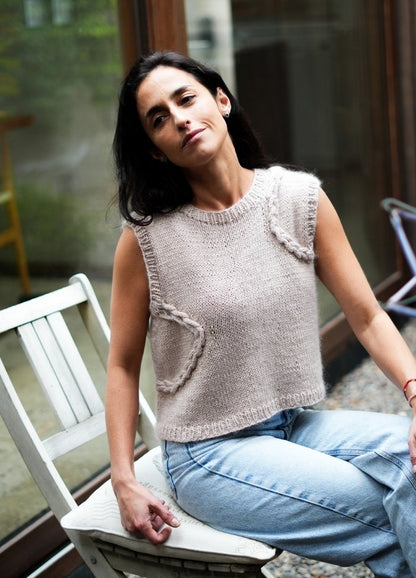 Woman with long dark hair wearing a beige knit sleeveless top and light blue jeans, sitting on a white chair by a window, exuding a relaxed and contemplative vibe.