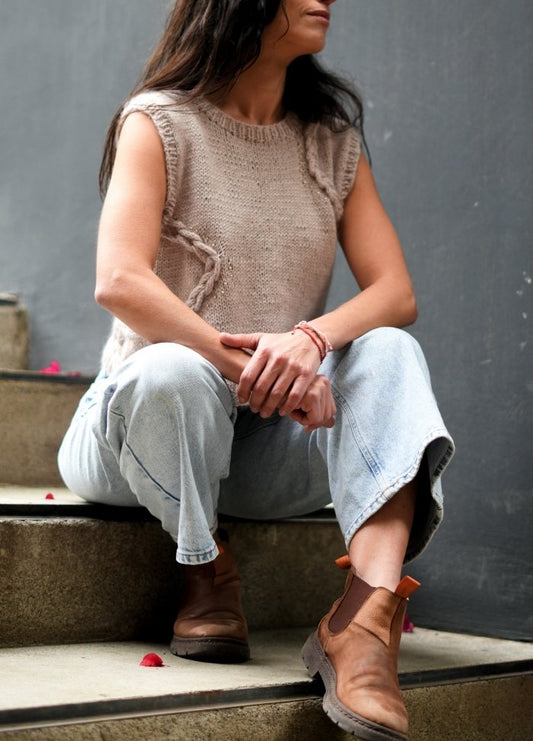 Woman sitting on stairs, wearing a sleeveless beige sweater and light blue jeans. Brown boots; relaxed pose. Scattered petals add a touch of color.