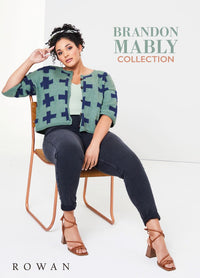 A woman sits confidently on a chair wearing a green cardigan with blue crosses, dark jeans, and strappy sandals. Text reads "Brandon Mably Collection." Calm, stylish ambiance.