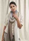 A person with dark hair, wearing a button-up cream blouse and long knitted scarf in beige and gray tones, stands against a light curtain backdrop, exuding elegance and calmness.