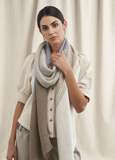 A person with dark hair, wearing a button-up cream blouse and long knitted scarf in beige and gray tones, stands against a light curtain backdrop, exuding elegance and calmness.