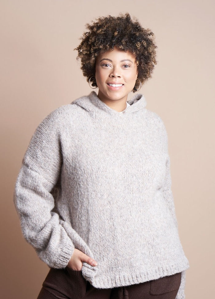 A person with curly hair smiles warmly, wearing a cozy light gray sweater with one hand in a pocket, set against a neutral beige background.