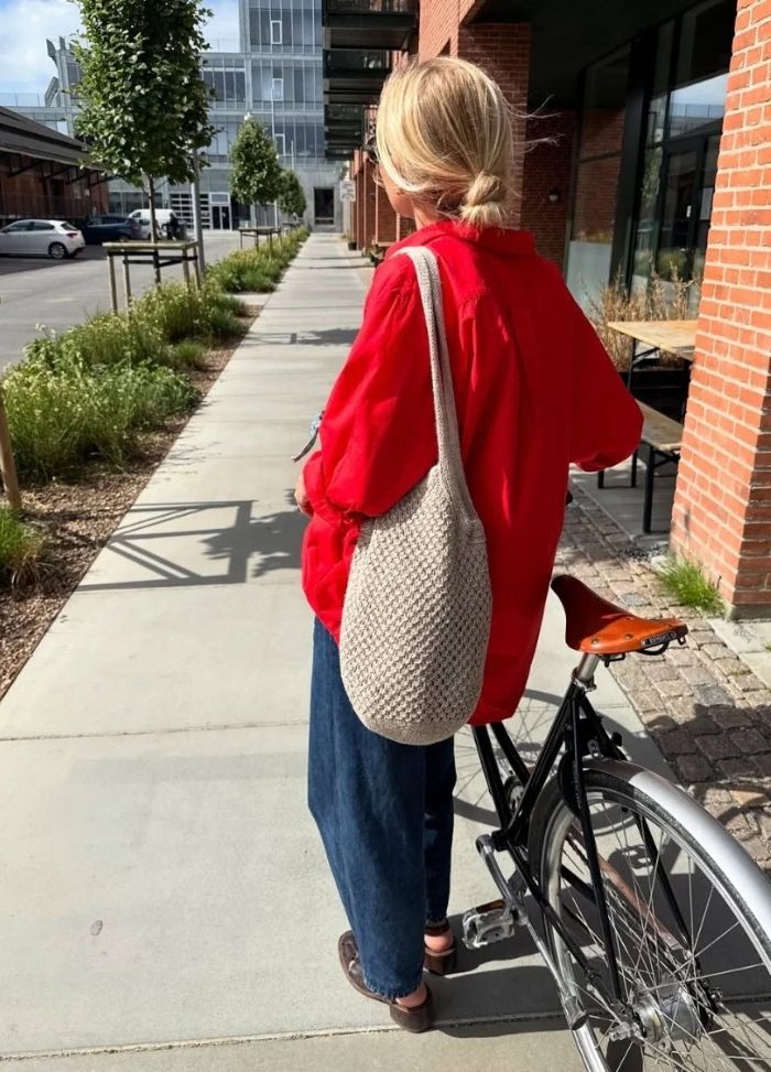 A person with blonde hair, wearing a red shirt and blue jeans, stands by a bicycle on a sunny sidewalk. They carry a beige woven bag.