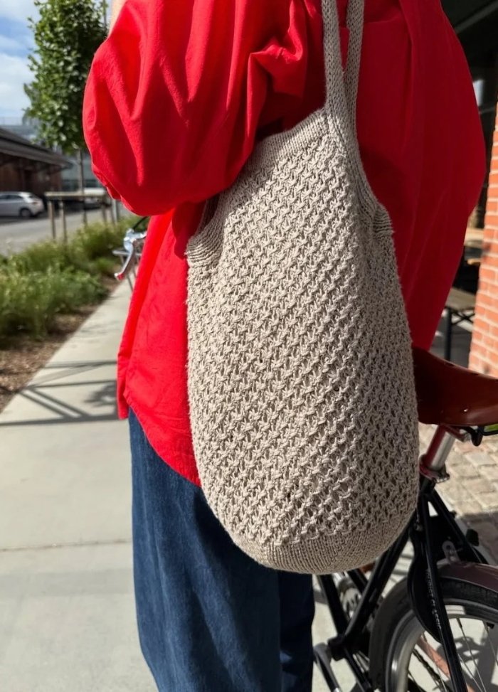 Person wearing a bright red top and blue jeans walks a bicycle on a sidewalk. They carry a textured beige knit bag over their shoulder.