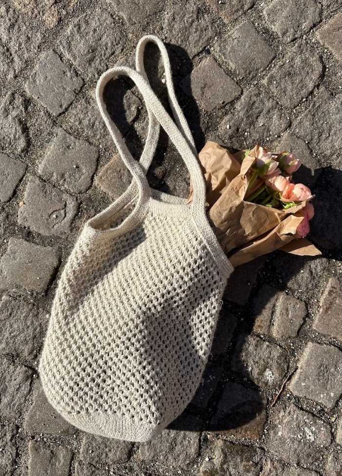 A white knitted bag lies on a sunlit cobblestone street, with pink flowers in brown paper peeking out, evoking a rustic, serene vibe.