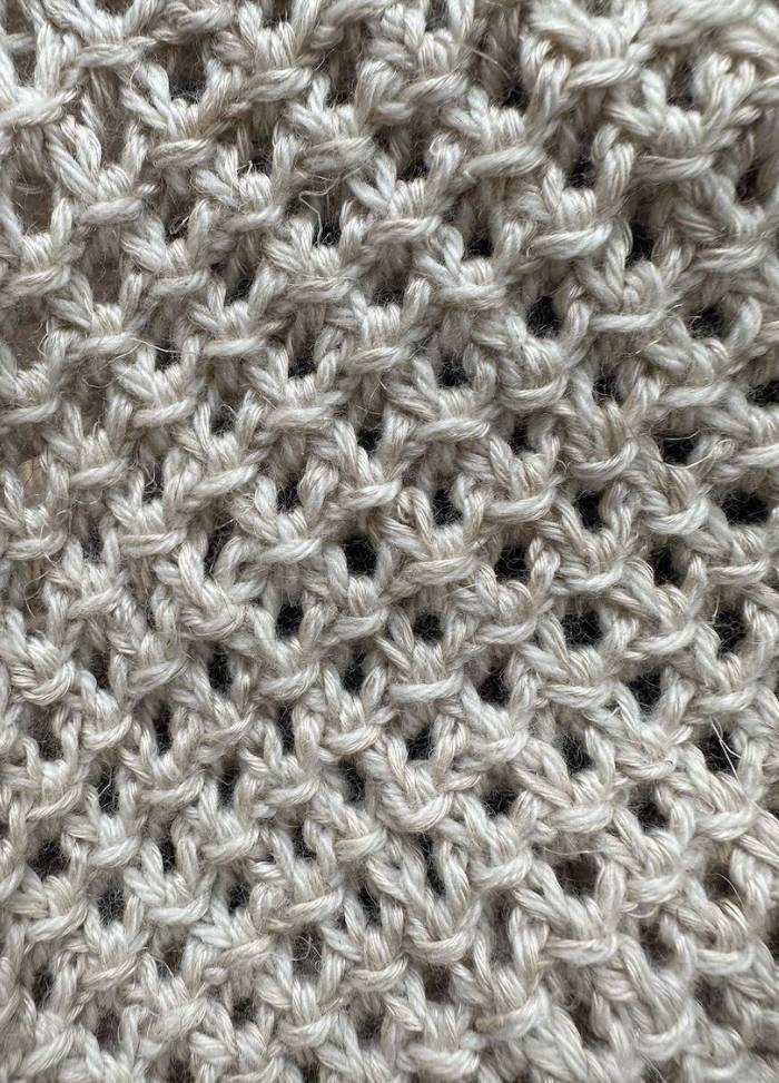 Close-up of beige knitted fabric with an open weave pattern, creating a textured, airy feel. The soft yarn conveys warmth and comfort.
