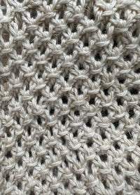 Close-up of beige knitted fabric with an open weave pattern, creating a textured, airy feel. The soft yarn conveys warmth and comfort.