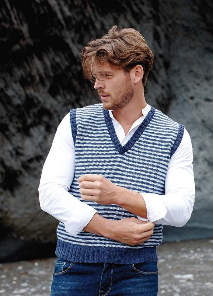 A man in a blue and white striped knit vest adjusts his white shirt sleeves. He stands on a rocky beach, gazing to the side with a thoughtful expression.