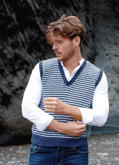 A man in a blue and white striped knit vest adjusts his white shirt sleeves. He stands on a rocky beach, gazing to the side with a thoughtful expression.