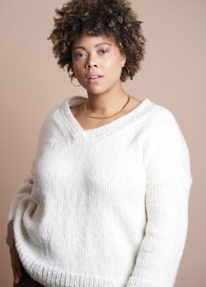 Woman with curly hair wearing a cozy, white sweater poses confidently against a soft, beige background. She has a calm and relaxed expression.