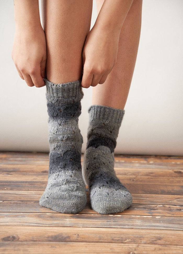 Close-up of a person wearing cozy, patterned wool socks with shades of gray and blue, standing on rustic wooden flooring, conveying warmth and comfort.
