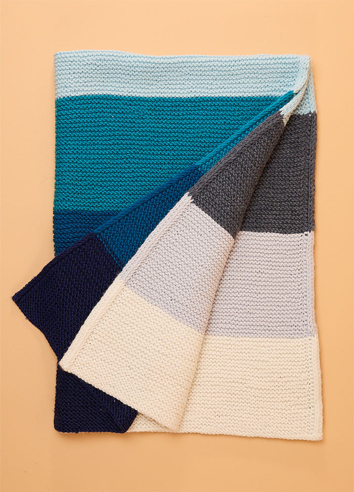 Folded hand-knitted Brighton Blanket with horizontal stripes in shades of blue, gray, and cream, showing both front and back sides against a light peach background – soft and serene color palette for cozy home decor.