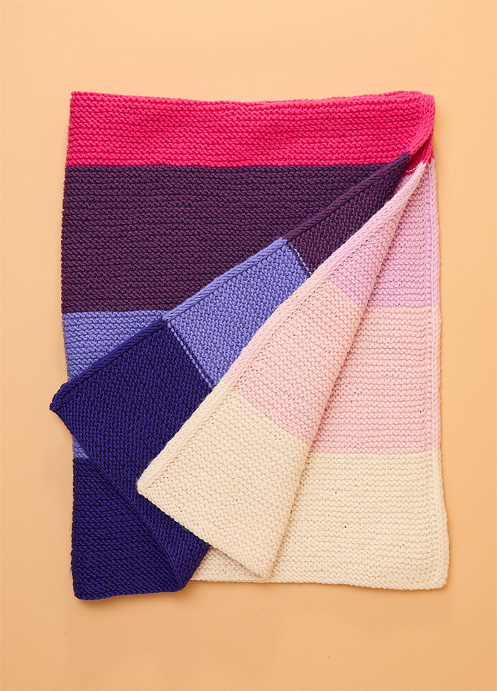Folded hand-knitted Brighton Blanket with horizontal stripes in bright pink, dark purple, light purple, blue, dark blue, cream, light pink, and white – colorful and cozy design showcasing a soft gradient palette.