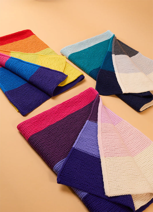 Three hand-knitted Brighton Blankets laid out on a beige surface – top left with rainbow stripes, top right in blue and gray tones, bottom left in pink, purple, and blue, and bottom right in soft pastel shades of white, pink, yellow, and light blue.