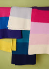 Hand-knitted Brighton Blanket with horizontal stripes in shades of pink, red, white, blue, purple, yellow, and green, displayed on a light green background – vibrant and cozy multicolor design.
