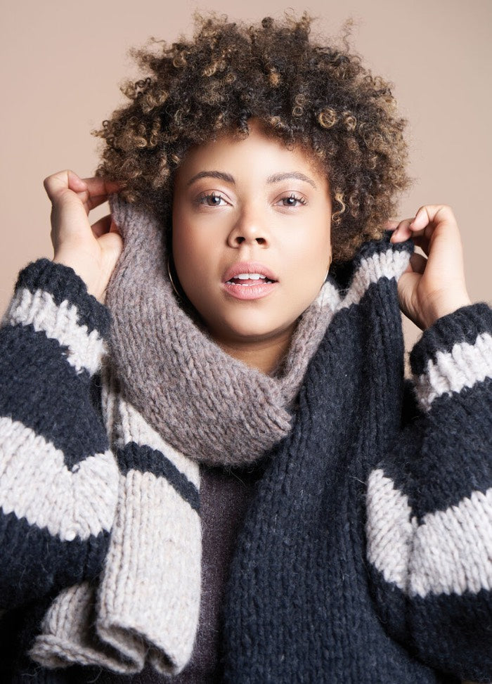 A person with curly hair, wearing a cozy, oversized striped sweater and chunky scarf, poses warmly against a neutral background, conveying a sense of comfort.