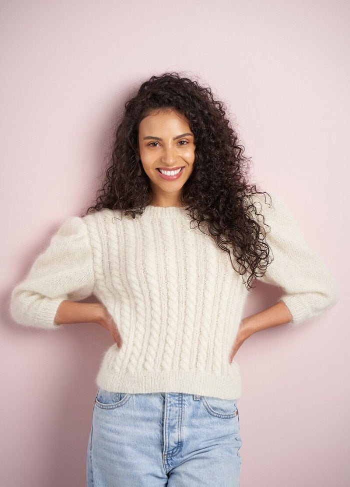 Smiling woman with curly hair wearing a cream cable-knit sweater and light blue jeans, standing against a soft pink background. The mood is cheerful.