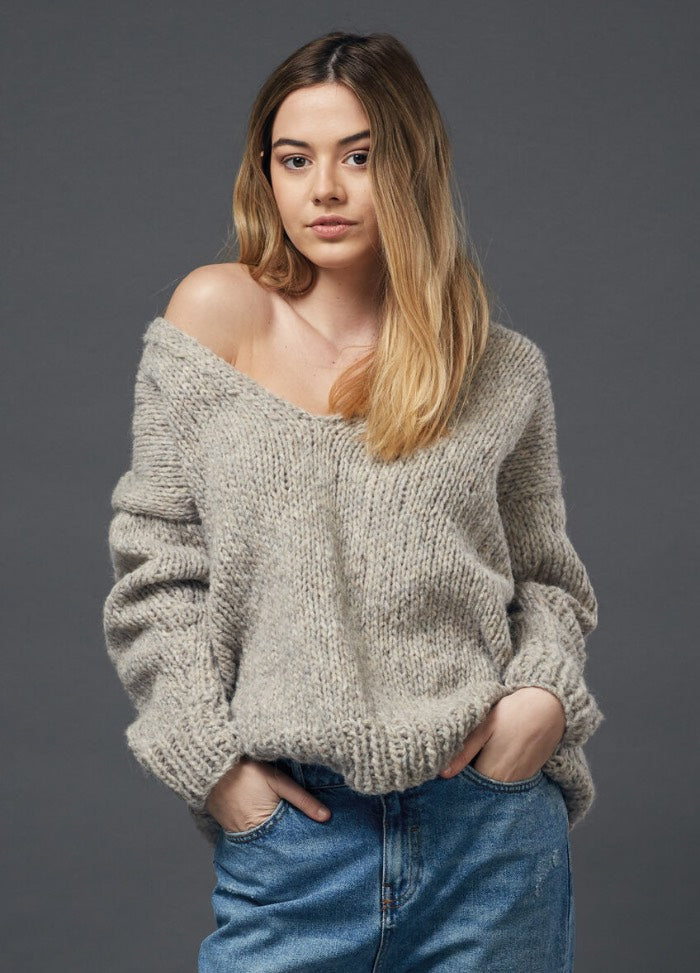 A woman with long hair wearing an oversized knitted gray sweater and blue jeans stands confidently against a plain gray background.