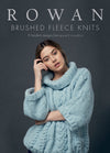 Model in a soft, thick blue sweater poses thoughtfully against a gray background. Text above reads: "Rowan Brushed Fleece Knits." Cozy and elegant.