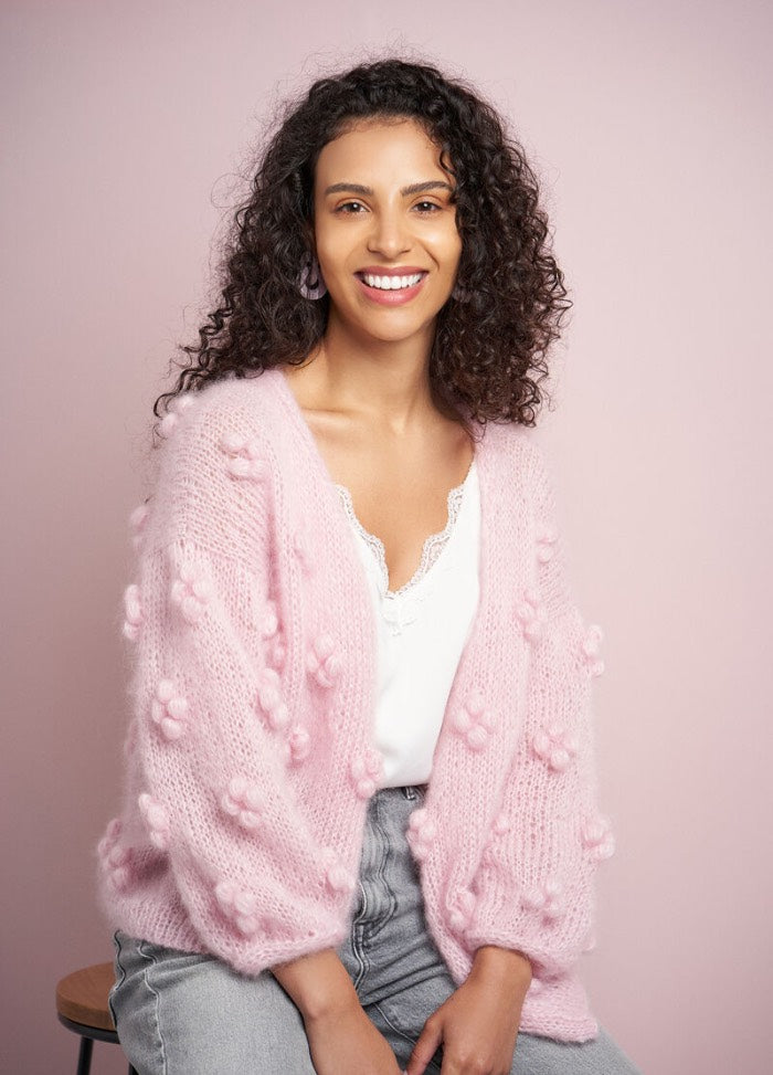 Smiling woman with curly hair wearing a pink fluffy cardigan with bobbles over a white top. She sits on a stool against a pink background.