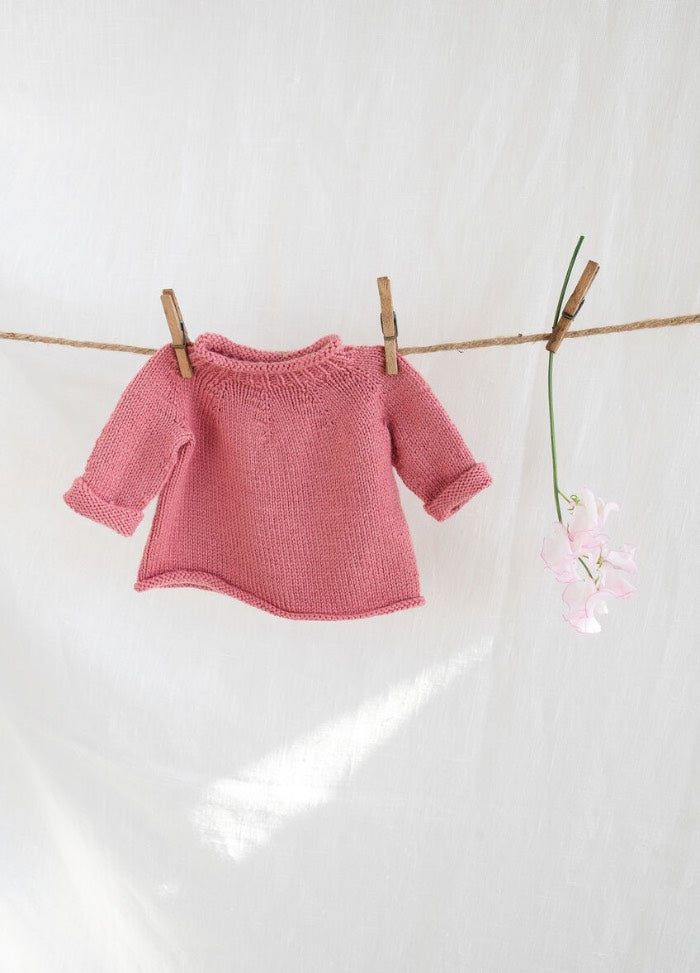 A pink knitted baby sweater hangs on a clothesline with clothespins. A delicate pink flower is attached, set against a soft white background.
