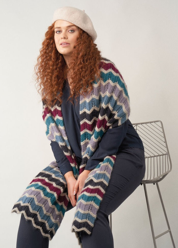 Long wrap scarf in shades of blue, grey, and cream, worked in bold zigzag chevrons with textured ridges, draped loosely over the shoulders.