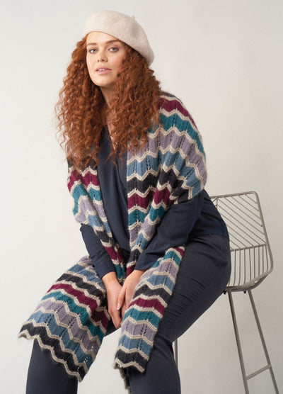 Long wrap scarf in shades of blue, grey, and cream, worked in bold zigzag chevrons with textured ridges, draped loosely over the shoulders.