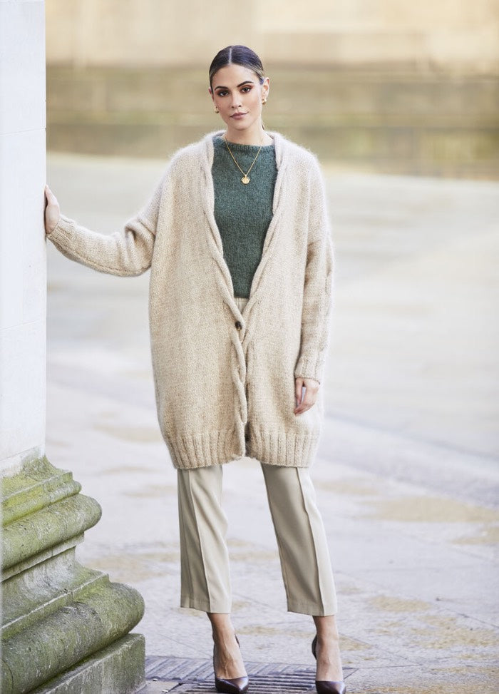 A woman leans against a stone wall, wearing a long beige cardigan over a green top and beige pants. The setting is an outdoor, urban environment.