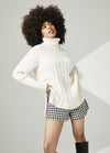 Woman with curly hair poses confidently in a white turtleneck sweater, plaid shorts, and black boots against a neutral background, conveying a stylish and cozy vibe.