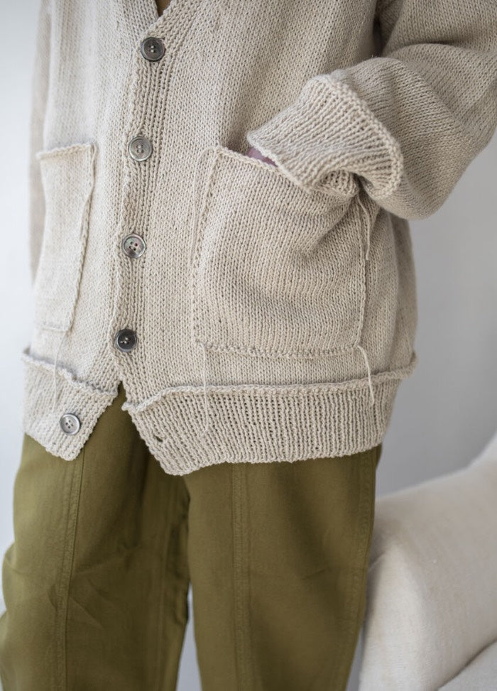Close-up of a person wearing a beige knit cardigan with buttons and two pockets, paired with olive green pants. The mood is relaxed and casual.